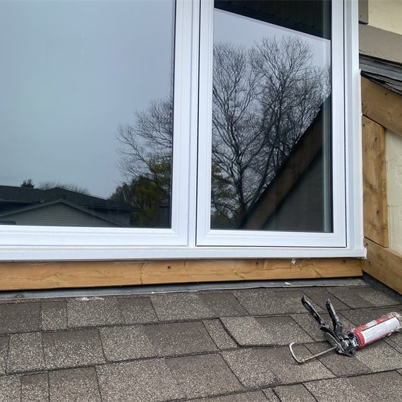 caulking gun resting on roof shingles in front of windows midway through window capping job